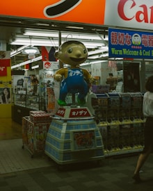 A whimsical cartoon character statue stands prominently outside a Japanese store. The scene is bustling with bright signage and vending machines filled with toys or collectibles. The interior reveals a glimpse of various products and advertisements, reflecting a vibrant commercial environment.