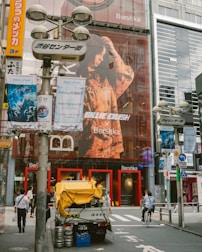 An urban street scene featuring a large storefront with a prominent advertisement of a person on the facade. Pedestrians and a cyclist are visible in the foreground. Several posters and signs with colorful graphics and text are seen on surrounding poles and buildings. A delivery truck with items like barrels and a yellow cover is parked on the street.