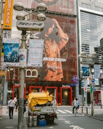 An urban street scene featuring a large storefront with a prominent advertisement of a person on the facade. Pedestrians and a cyclist are visible in the foreground. Several posters and signs with colorful graphics and text are seen on surrounding poles and buildings. A delivery truck with items like barrels and a yellow cover is parked on the street.