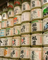 A collection of traditional Japanese sake barrels is stacked together, featuring a variety of designs and calligraphy in vibrant colors including red, black, and green. The barrels appear to be made of a woven material and are ornately decorated, symbolizing cultural or ceremonial significance.