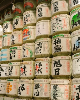 A collection of traditional Japanese sake barrels is stacked together, featuring a variety of designs and calligraphy in vibrant colors including red, black, and green. The barrels appear to be made of a woven material and are ornately decorated, symbolizing cultural or ceremonial significance.