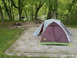 A camping scene with a pitched tent on a gravel area surrounded by lush green trees. Nearby, there is a picnic table and a grill next to a fire pit, which has some logs placed around it for sitting. The environment is serene with a relaxed outdoor vibe.