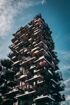 A modern skyscraper with balconies filled with lush trees and plants, creating a vertical forest effect. The building's architecture features staggered terraces, presenting a mix of greenery and concrete against a blue sky with scattered clouds.