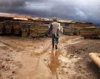A person wearing work clothes and a helmet walks through a muddy area with mining carts on either side. The sky is overcast with dark, stormy clouds, and the ground is covered in brown mud.