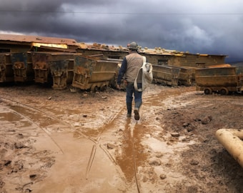 A person wearing work clothes and a helmet walks through a muddy area with mining carts on either side. The sky is overcast with dark, stormy clouds, and the ground is covered in brown mud.