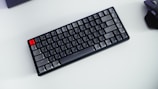 A compact mechanical keyboard with gray, black, and one red key placed on a light background. A wireless mouse is partially visible on the right side.