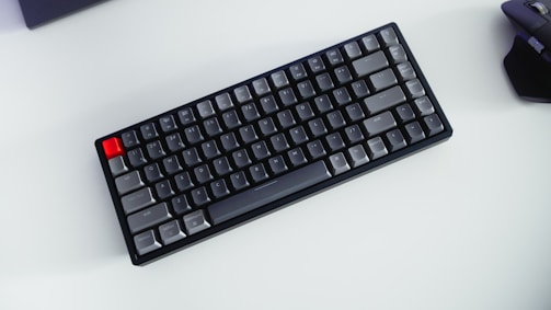 A compact mechanical keyboard with gray, black, and one red key placed on a light background. A wireless mouse is partially visible on the right side.