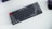 A compact mechanical keyboard with gray, black, and one red key placed on a light background. A wireless mouse is partially visible on the right side.