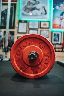 A large red weight plate labeled 'YORK 45 LB' is prominently placed on a gym floor. In the background, the walls are adorned with framed pictures and posters, some of which have a neon green border.