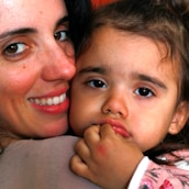 A close-up of a woman smiling as she embraces a young child. The child has a thoughtful expression and rests their hand near their mouth. Both have dark hair and eyes.