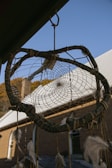 A dreamcatcher with a woven web and dangling feathers hangs outside under an eave. The background features a clear blue sky and a building with white roofs and brick walls.