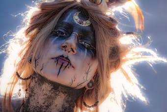 A person with elaborate makeup featuring black and white paint, highlighted by a crescent moon decoration on the forehead. The hair is styled dramatically with light shining through, creating a halo effect. Several facial piercings and dark eyeliner contribute to an otherworldly appearance. Intricate patterns are drawn across the face and neck, accentuating the mystical theme.