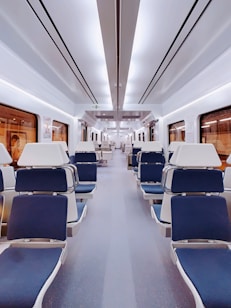 An empty, modern train interior with comfortable blue and white padded seats arranged in rows on either side of a central aisle. The lighting is bright and evenly spread across the ceiling, with large windows on the sides of the cabin.