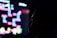 A silhouette of a person is seen in front of a brightly colored, pixelated screen. The screen displays a retro-style video game with vibrant geometric shapes in neon pinks, blues, and yellows. The background is dark, highlighting the contrast between the dark silhouette and the luminous screen.