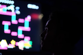 A silhouette of a person is seen in front of a brightly colored, pixelated screen. The screen displays a retro-style video game with vibrant geometric shapes in neon pinks, blues, and yellows. The background is dark, highlighting the contrast between the dark silhouette and the luminous screen.