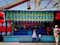 A carnival game booth is adorned with various stuffed toys including numerous blue turtle-like creatures, red and pink creatures, and a yellow mouse-like figure with pointed ears. The booth is part of a larger amusement area with visible roller coaster tracks above. Two children stand at the booth's front near several targets, accompanied by an adult who appears to be engaging with a staff member in a red uniform.