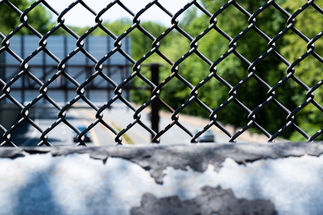 A chain-link fence with a blurry view of a highway and green trees in the background. The metal fence is prominently in focus, while the road and natural scenery beyond appear out of focus.