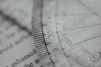 A close-up view of a transparent protractor laid over a page with typed text. The focus is on the markings on the protractor, including numbers and degree measurements, while the background text is blurred.