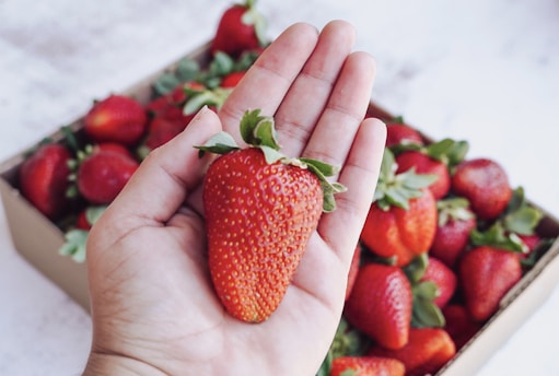 A hand holding a large, ripe strawberry above a box full of strawberries. The berries are fresh, vivid red with green leaves, and the scene captures the freshness and natural appeal of the fruit.