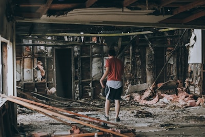 A person wearing a red shirt and shorts is standing in the middle of a destroyed building with exposed insulation and wooden supports. The surroundings are cluttered with debris, broken structures, and insulation material scattered on the floor.