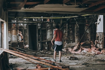 A person wearing a red shirt and shorts is standing in the middle of a destroyed building with exposed insulation and wooden supports. The surroundings are cluttered with debris, broken structures, and insulation material scattered on the floor.