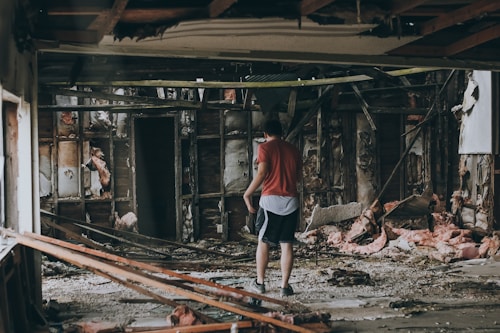 A person wearing a red shirt and shorts is standing in the middle of a destroyed building with exposed insulation and wooden supports. The surroundings are cluttered with debris, broken structures, and insulation material scattered on the floor.