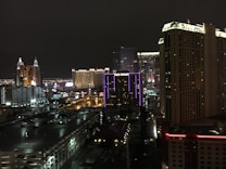 A nighttime cityscape showcasing a vibrant urban skyline with illuminated high-rise buildings. Neon lights and colorful signage are prominently visible, contributing to the lively atmosphere. Streets and parking lots are seen below the towering structures.