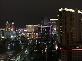 A nighttime cityscape showcasing a vibrant urban skyline with illuminated high-rise buildings. Neon lights and colorful signage are prominently visible, contributing to the lively atmosphere. Streets and parking lots are seen below the towering structures.