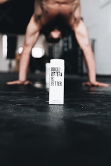 A person performs a handstand in the background while a carton of boxed water is prominently placed on the ground in the foreground. The carton has the text 'Boxed Water is Better' printed on it. The setting appears to be indoors, possibly a gym or studio, with a focus on fitness and sustainability.