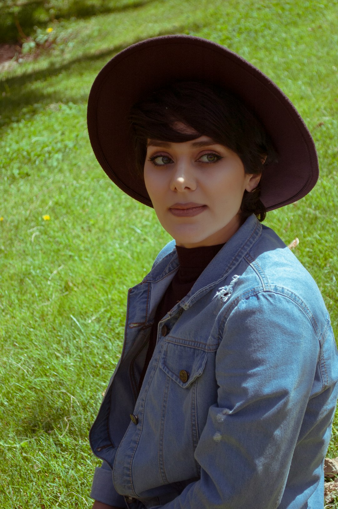girl in blue denim jacket and brown hat sitting on green grass field during daytime