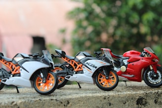 Three miniature sport motorcycles are positioned on a rough surface. Two of the bikes are white with orange details, while the third is red. The background is blurred with greenery.