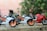 Three miniature sport motorcycles are positioned on a rough surface. Two of the bikes are white with orange details, while the third is red. The background is blurred with greenery.