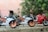 Three miniature sport motorcycles are positioned on a rough surface. Two of the bikes are white with orange details, while the third is red. The background is blurred with greenery.