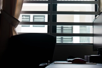 A dark office setting with a window revealing light from outside. Indoor elements include a computer monitor, a wireless mouse, and an office chair mostly in silhouette. The window blinds are partially open, allowing some light to illuminate the space.