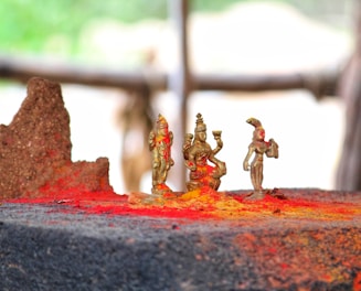 Small brass statues surrounded by vibrant red and yellow powders, likely representing traditional religious figures, placed on a dark stone surface with a blurred natural background.