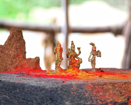 Small brass statues surrounded by vibrant red and yellow powders, likely representing traditional religious figures, placed on a dark stone surface with a blurred natural background.