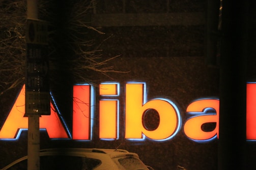 A brightly illuminated red and blue sign against a dark background. The letters 'Ali' are visible, with the rest obscured. Nearby, a tree with bare branches and a signpost are visible.