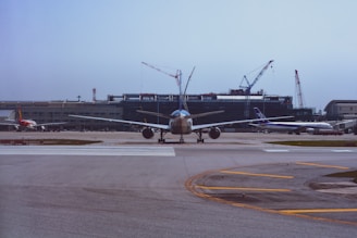 A large commercial airplane is positioned on a runway, ready for takeoff at an airport. The background features a series of cranes and a terminal building under construction. Additional aircraft are parked on the tarmac near the terminal.