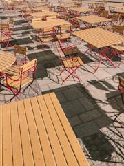 Outdoor seating arrangement with multiple wooden tables and chairs featuring a bright red metal frame. The furniture is placed on a paved surface, casting distinct shadows on the ground.