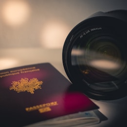 A close-up of a passport with a European emblem partially visible, resting next to a camera lens. The passport cover is dark red, and the camera lens has prominent branding. The focus creates a soft, blurred background.