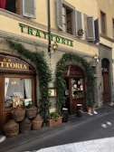 The exterior of an Italian trattoria with a rustic and traditional appearance. Ivy climbs over stone walls and surrounds large wooden doors and windows. Several wicker baskets and potted plants are placed along the sidewalk near barrels. The building features green shutters and a vintage street lamp beside a sign reading 'TRATTORIA' in green letters.