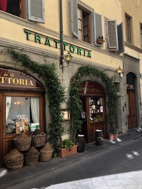 The exterior of an Italian trattoria with a rustic and traditional appearance. Ivy climbs over stone walls and surrounds large wooden doors and windows. Several wicker baskets and potted plants are placed along the sidewalk near barrels. The building features green shutters and a vintage street lamp beside a sign reading 'TRATTORIA' in green letters.
