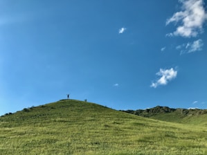 A person stands triumphantly at the peak of a gently sloping, grassy hill under a vast, clear blue sky with a few scattered clouds.