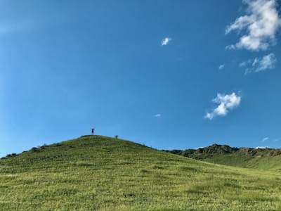 A person stands triumphantly at the peak of a gently sloping, grassy hill under a vast, clear blue sky with a few scattered clouds.