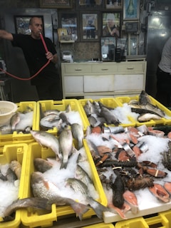 A man is standing in a shop, holding a smoking hookah pipe. In the foreground, yellow crates are filled with fresh fish, salmon steaks, and lobsters displayed on ice. The shop has a rustic appearance with various framed photos and a few appliances visible in the background.