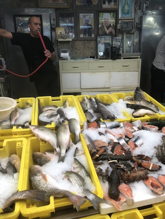 A man is standing in a shop, holding a smoking hookah pipe. In the foreground, yellow crates are filled with fresh fish, salmon steaks, and lobsters displayed on ice. The shop has a rustic appearance with various framed photos and a few appliances visible in the background.