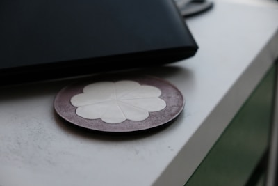 A round coaster with a floral design lies on a smooth surface next to a closed, dark-colored laptop or notebook. The coaster has a petal-like white pattern on a muted brown background.