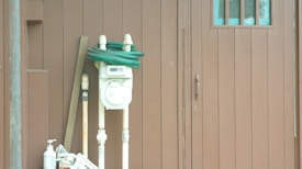 An exterior view of a brown wooden wall with three small window panes. A utility meter attached to the wall is surrounded by a coiled green hose, wooden sticks, and a bottle of liquid soap.