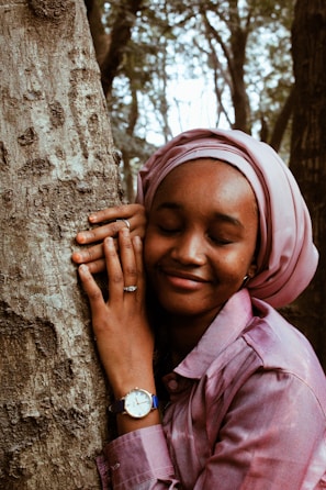 A person with a serene expression is embracing a tree trunk, wearing a pink headscarf and shirt. The watch and ring on their left hand suggest a sense of personal style. The background features a forest setting, with trees and diffused light creating a peaceful ambiance.