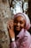 A person with a serene expression is embracing a tree trunk, wearing a pink headscarf and shirt. The watch and ring on their left hand suggest a sense of personal style. The background features a forest setting, with trees and diffused light creating a peaceful ambiance.